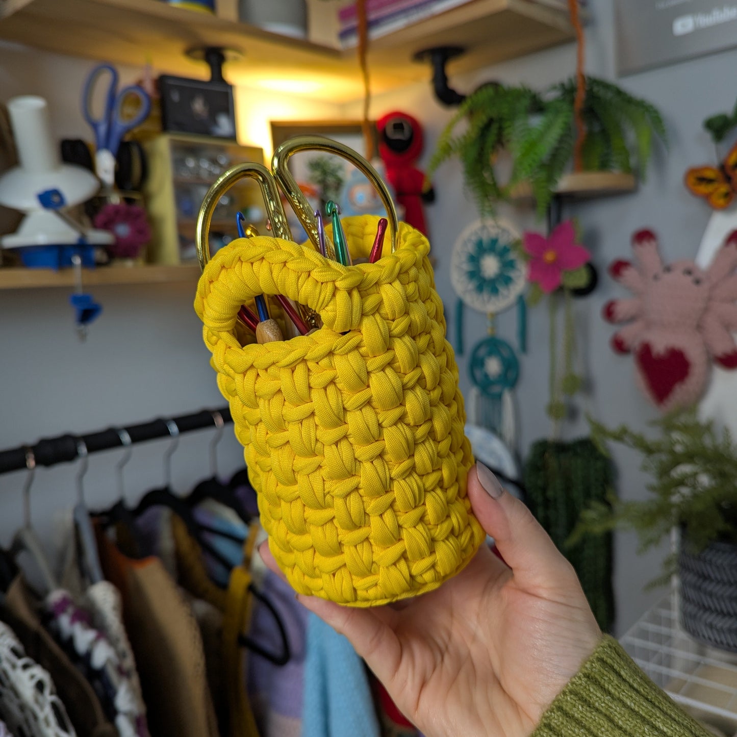 Yellow woven basket with pens held in front of a colorful background with shelves and plants.